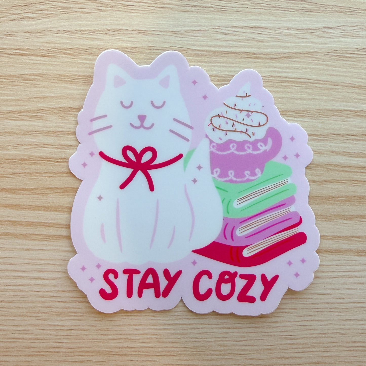 Sticker of a cat with books and hot drink and 'Stay Cozy' text on a wooden surface
