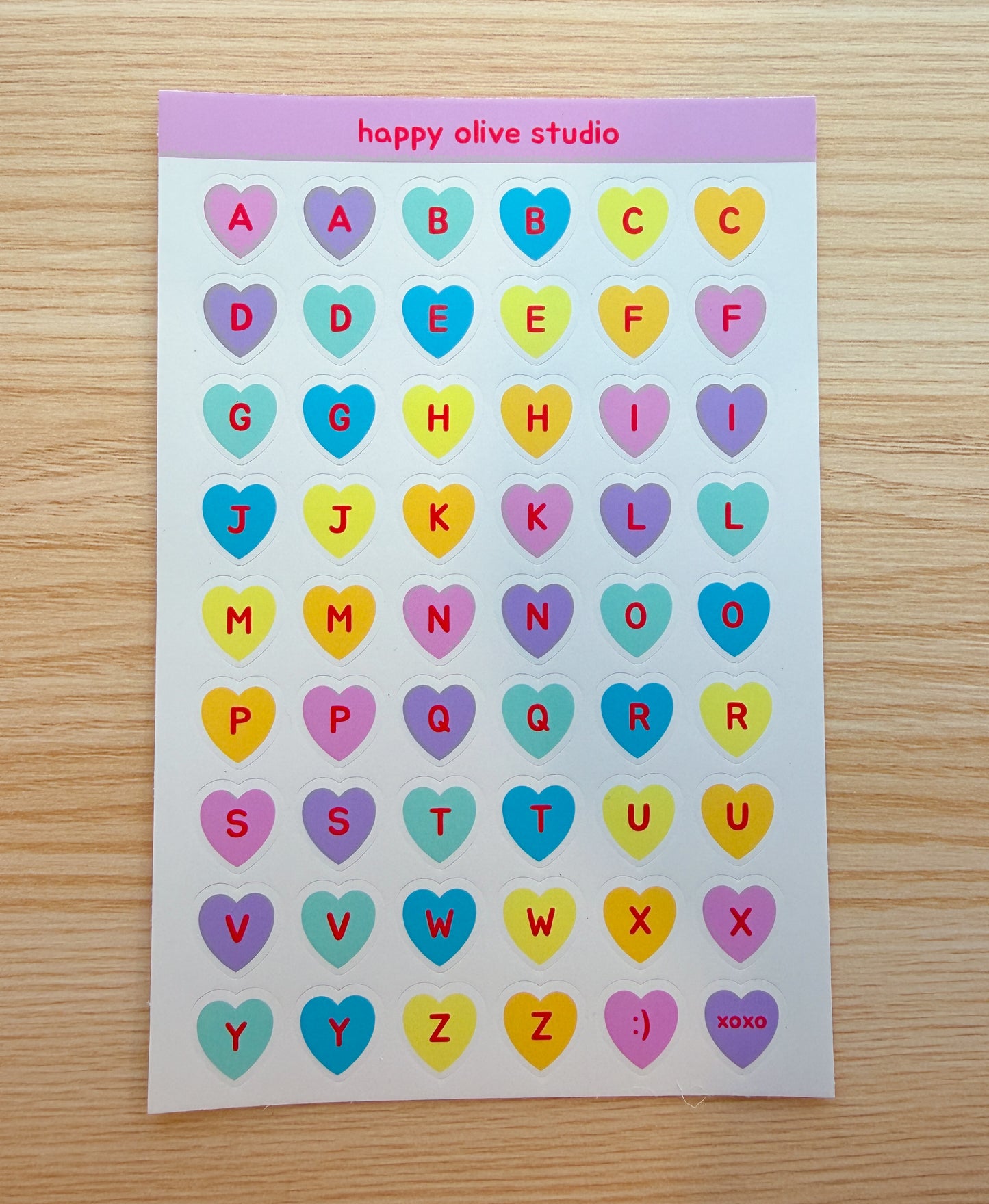 Sheet of colorful heart-shaped letter stickers with 'happy olive studio' branding on a wooden surface.