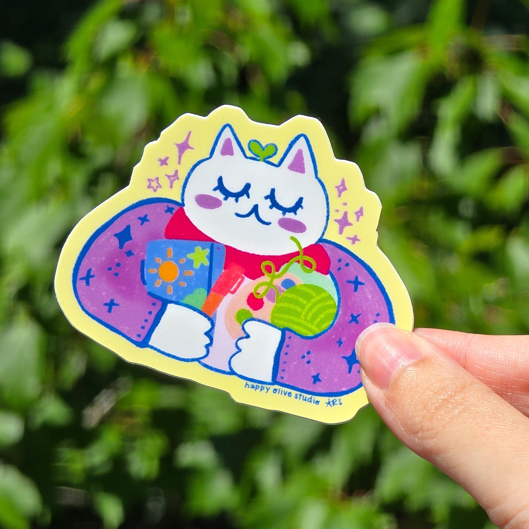 sticker of a cat holding various analog hobbies such as a journal, yarn, paint palette, and marker.