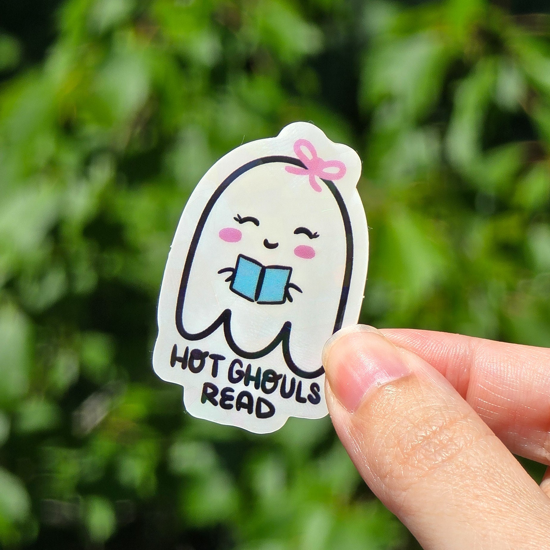sticker of a little white ghost, with a pink bow on her head holding a blue book. it says hot ghouls read. This sticker has a cracked ice holographic finish