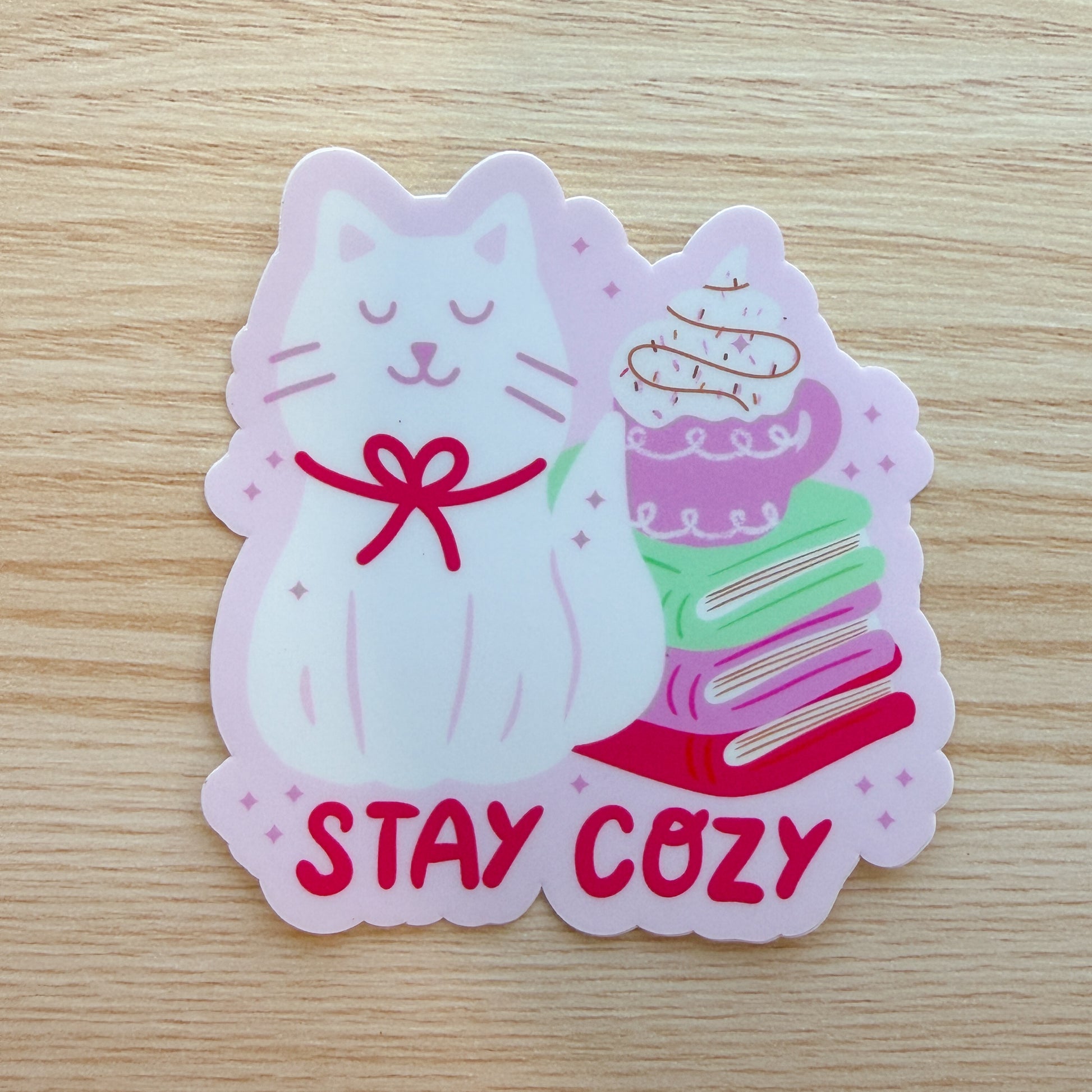 Sticker of a cat with books and hot drink and 'Stay Cozy' text on a wooden surface