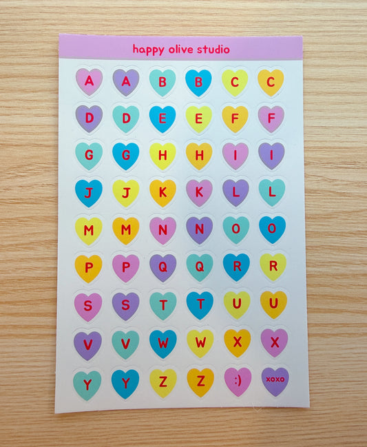 Sheet of colorful heart-shaped letter stickers with 'happy olive studio' branding on a wooden surface.